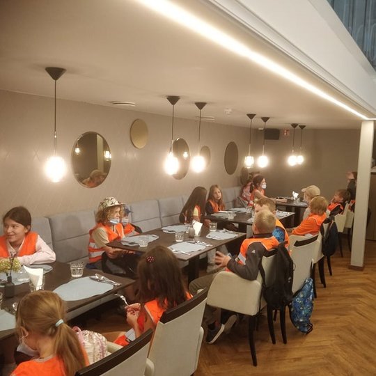 Vaikai pietauja kavinėje. Jie laukia maisto. Visi sėdi prie stalų, kurie yra paserviruoti.