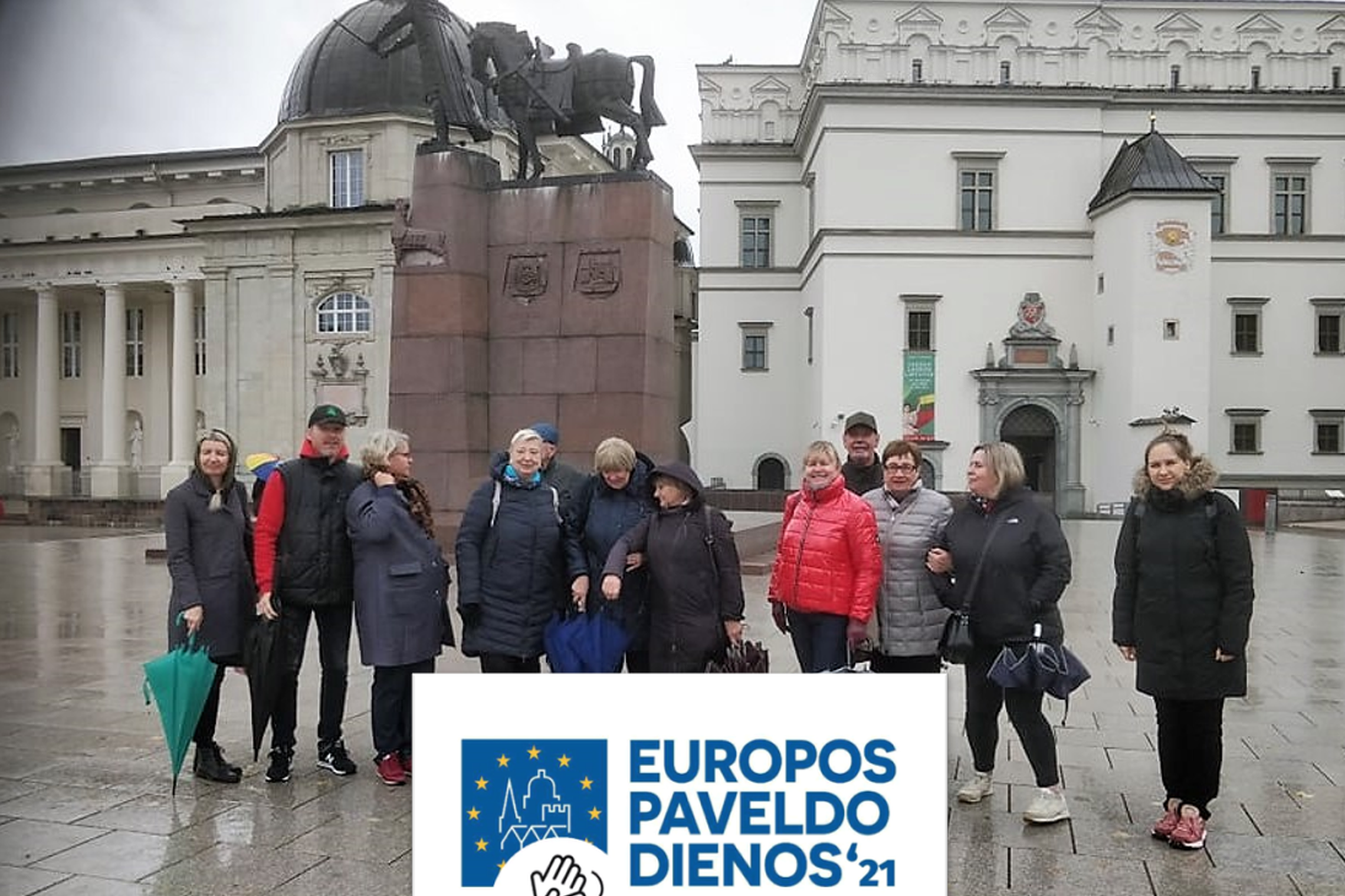 Ekskursijos dalyviai stovi Katedros aikštėje netoli Gedimino paminklo. Už jų nugarų, yra Valdovų rūmai ir Vilniaus katedra. Ką tik pasibaigė lietus, nes žmonės turi nesuglaustus skėčius. Dangus pilkas, be saulės. Ant nuotraukos užrašyta EUROPOS PAVELDO D