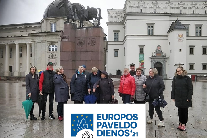 Ekskursijos dalyviai stovi Katedros aikštėje netoli Gedimino paminklo. Už jų nugarų, yra Valdovų rūmai ir Vilniaus katedra. Ką tik pasibaigė lietus, nes žmonės turi nesuglaustus skėčius. Dangus pilkas, be saulės. Ant nuotraukos užrašyta EUROPOS PAVELDO D