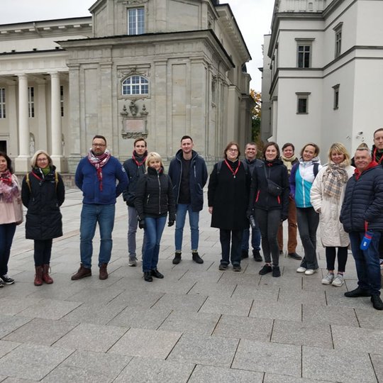 Ekskursijos dalyviai stovi Katedros aikštėje netoli Gedimino paminklo. Grupė yra didelė. Už jų nugarų, yra Valdovų rūmai ir Vilniaus katedra.