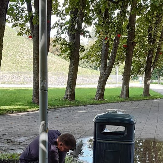 Jaunas vyriškis, apsirengęs tamsiai pilkais, trumpom rankovėm marškinėliais, tokios pat spalvos kelnėm, storapadžiais batais, su darbinėm pirštinėm laiko žarną su „pistoletu”. Didelio spaudimo vandens srovė nutaikyta į šiukšliadėžės paviršių, srovė tokia 
