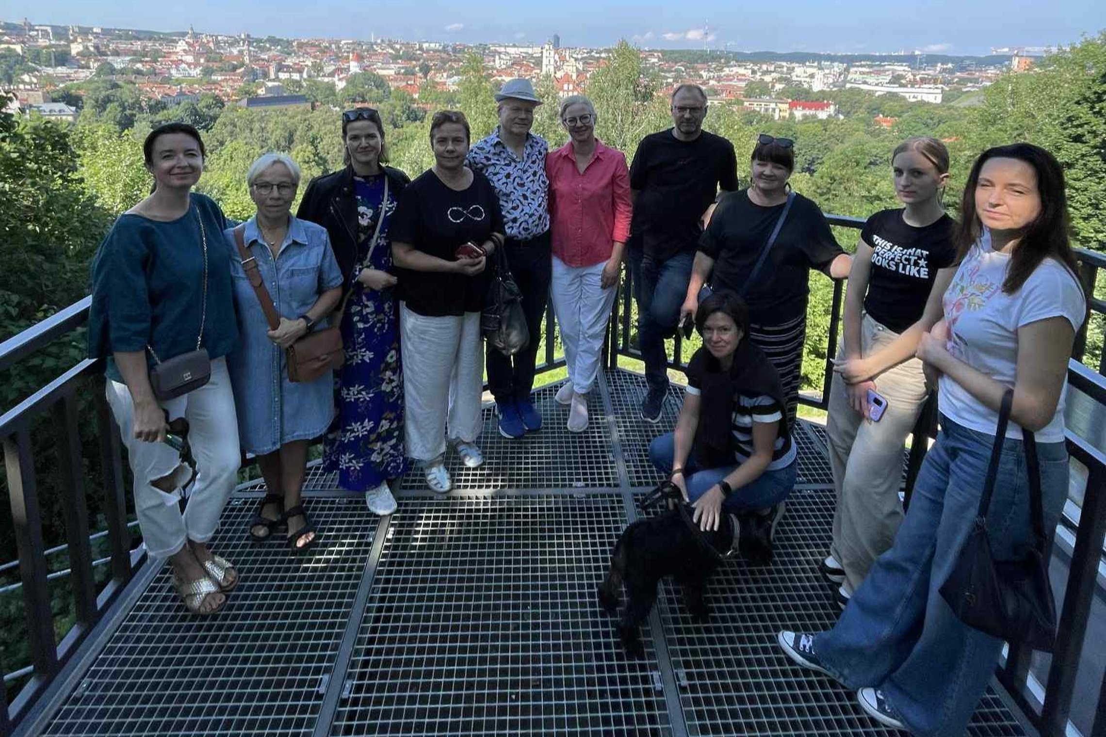 Grupė žmonių stovi ant Vilniaus pilių rezervato Alnatos kalno bokštelio apžvalgos aikštelės, kur atsiveria Vilniaus panorama – raudoni stogai, bažnyčių bokštai. Priekyje moteris glosto juodą šunį. Saulėta diena, visi atsipalaidavę ir šypsosi.