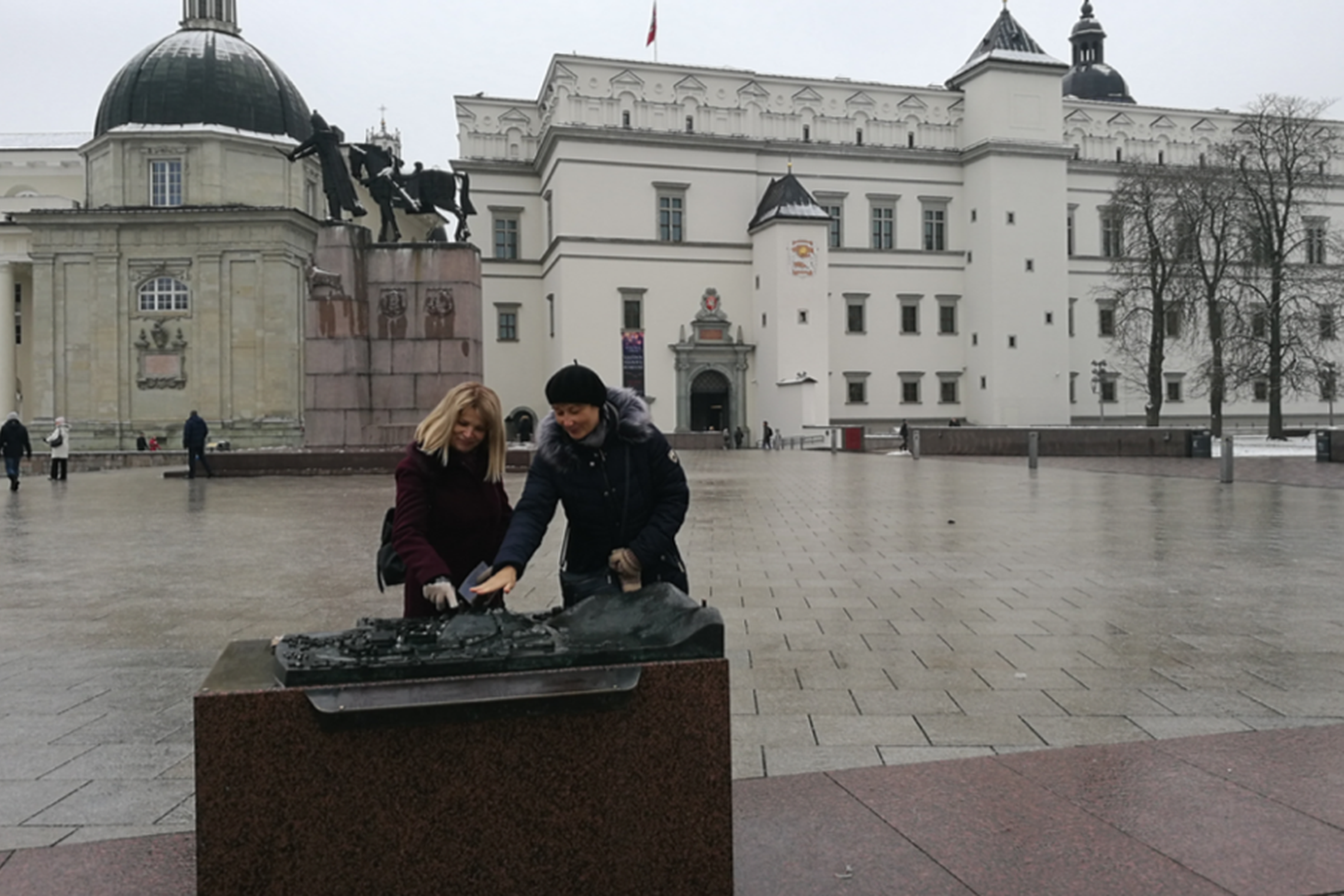Dvi moterys apžiūrinėja bronzinį Vilniaus pilių maketą Katedros aikštėje. Už jų nugaros stūkso Valdovų rūmai ir Gedimino paminklas. Kairėje Vilniaus katedros Šv. Kazimiero koplyčia.