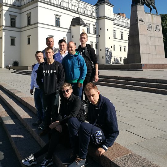 Grupė ekskursijos dalyvių stovi Valdovų rūmų ir Gedimino paminklo fone.