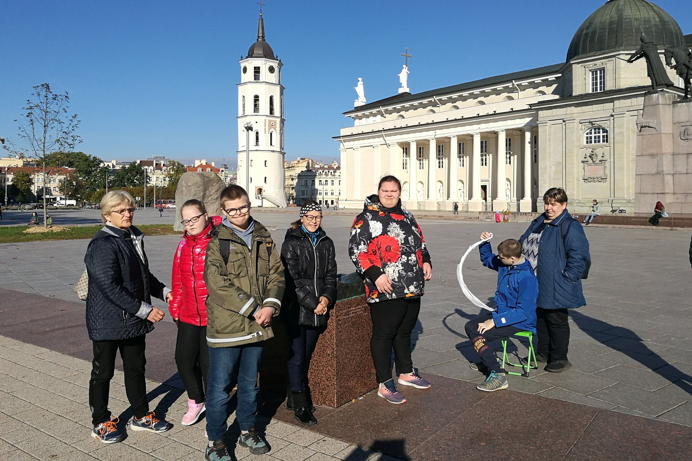  Penki vaikai ir dvi suaugusios moterys stovi prie metalinio Vilniaus pilių maketo Kateros aikštėje. Vaikai apsivilkę sriukes ir beveik visi yra su akinukais. Nuotraukos centre matosi Vilniaus katedros varpinė ir Katedra. Graži saulėta diena, danguje nei 