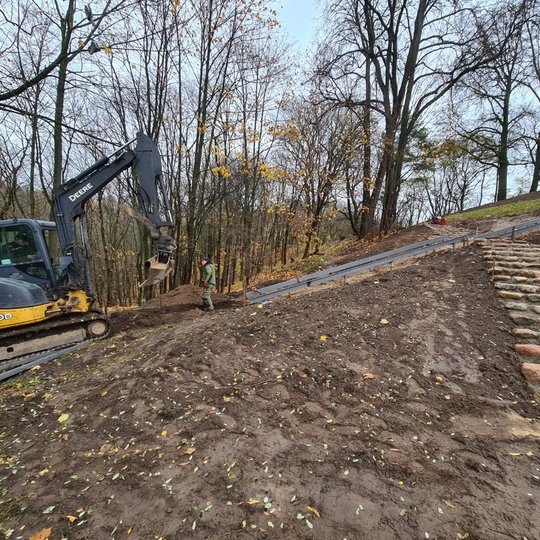 Pirmame trečdalyje nuotraukos yra juoda žemė. Įkalnė kyla į dešinę, taip įstrižai. Dešinėje randasi pradėtų mūryti laiptų konstrukcija: laiptai lipinami iš akmenų ir cemento mišinio. Akmenys nemaži, vienoje pakopoje yra 5 – 6 akmenys. Laiptų konstrukcija 