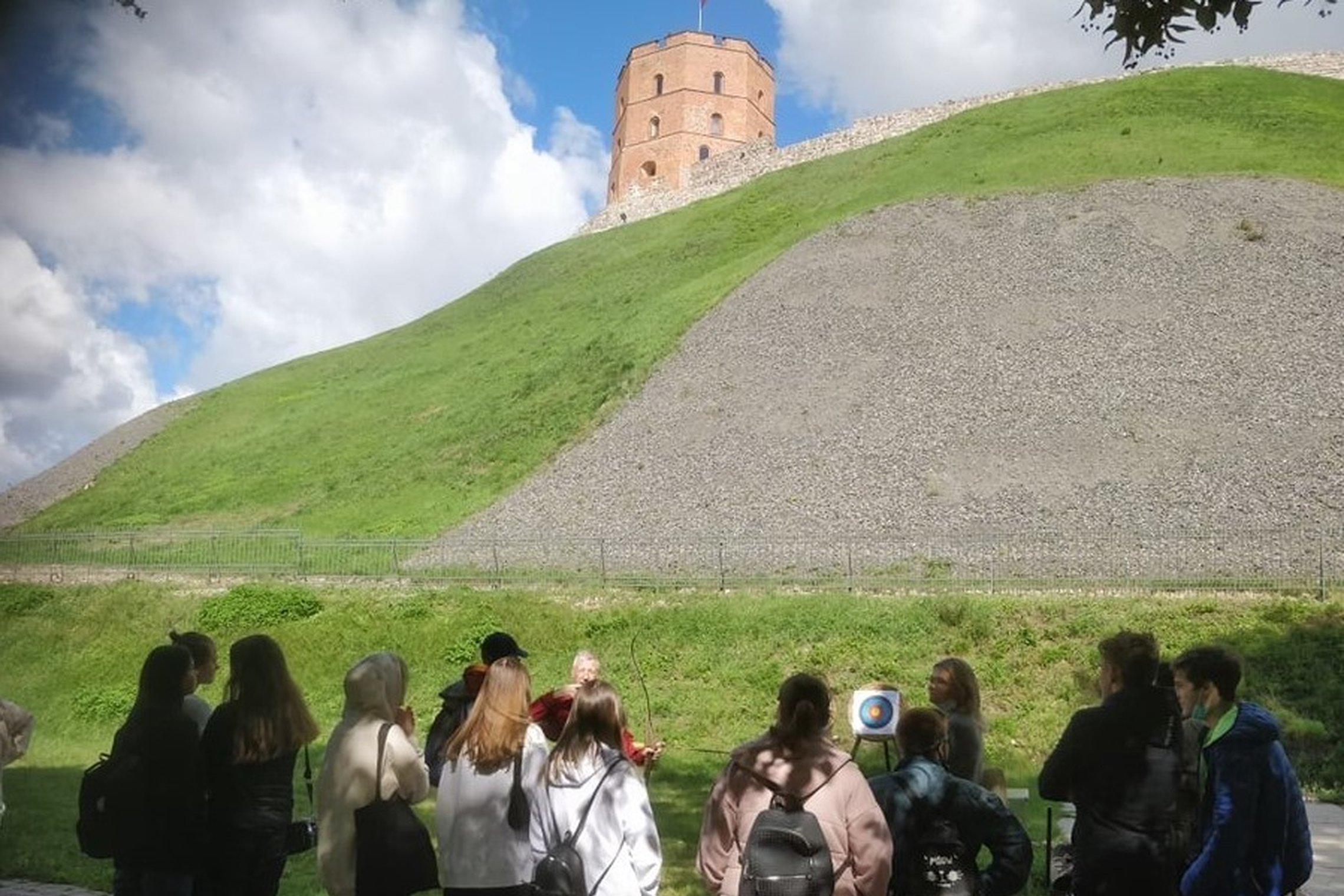 Nemaža grupė paauglių mokinių stovi Gedimino pilies kalno papėdėje ir šaudo iš lankų į taikinius. Matosi žydras su debesimis dangus.