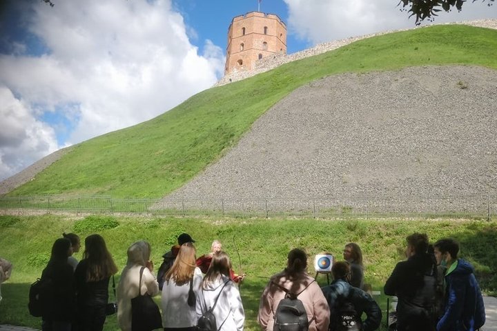 Nemaža grupė paauglių mokinių stovi Gedimino pilies kalno papėdėje ir šaudo iš lankų į taikinius. Matosi žydras su debesimis dangus.