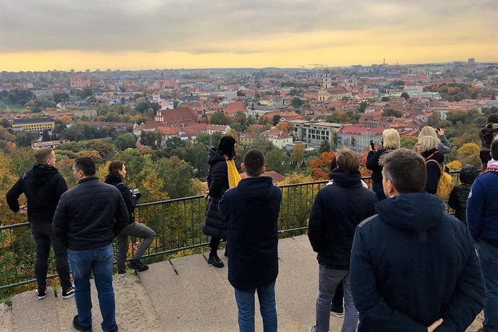Stovi grupė žmonių ant Trijų Kryžių kalno. Jie žiūri į miesto vaizdą, kuris nuo šio kalno atsiveria labai plačiai. Priekyje stovi ekskursijos gidė.