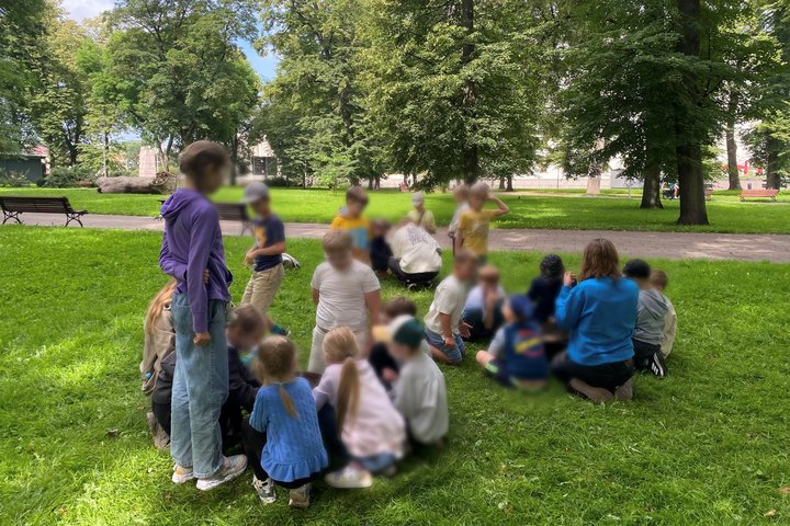 Vaikai susėdę ratu ant žolės parke, bando užkurti ugnį laužavietėse, vyksta edukacinis užsiėmimas