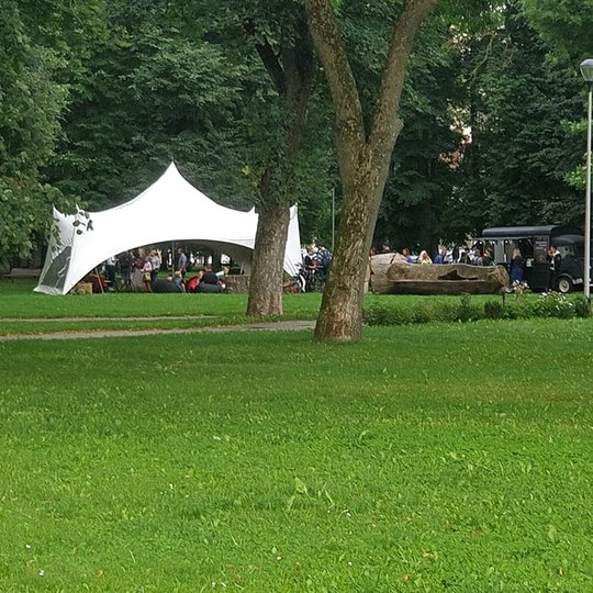 Parke ant žalios žolės, po medžiais pastatyta graži balta palapinė. Jo ja susėdę nemažai žmonių. Jie ten klausosi paskaitos. Netoli nuo palapinės stovi maisto autobusiukas, jame kavinės virtuvės įefas ir jis dalina maistą.