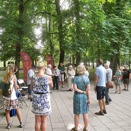 Pilių parke vyksta Vilniaus pilių paukščių nuotraukų parodos atidarymas. Susirinkę būrelis žmonių. Yra išdėliotos kėdės ir stalas. Aplinkui seni medžiai.