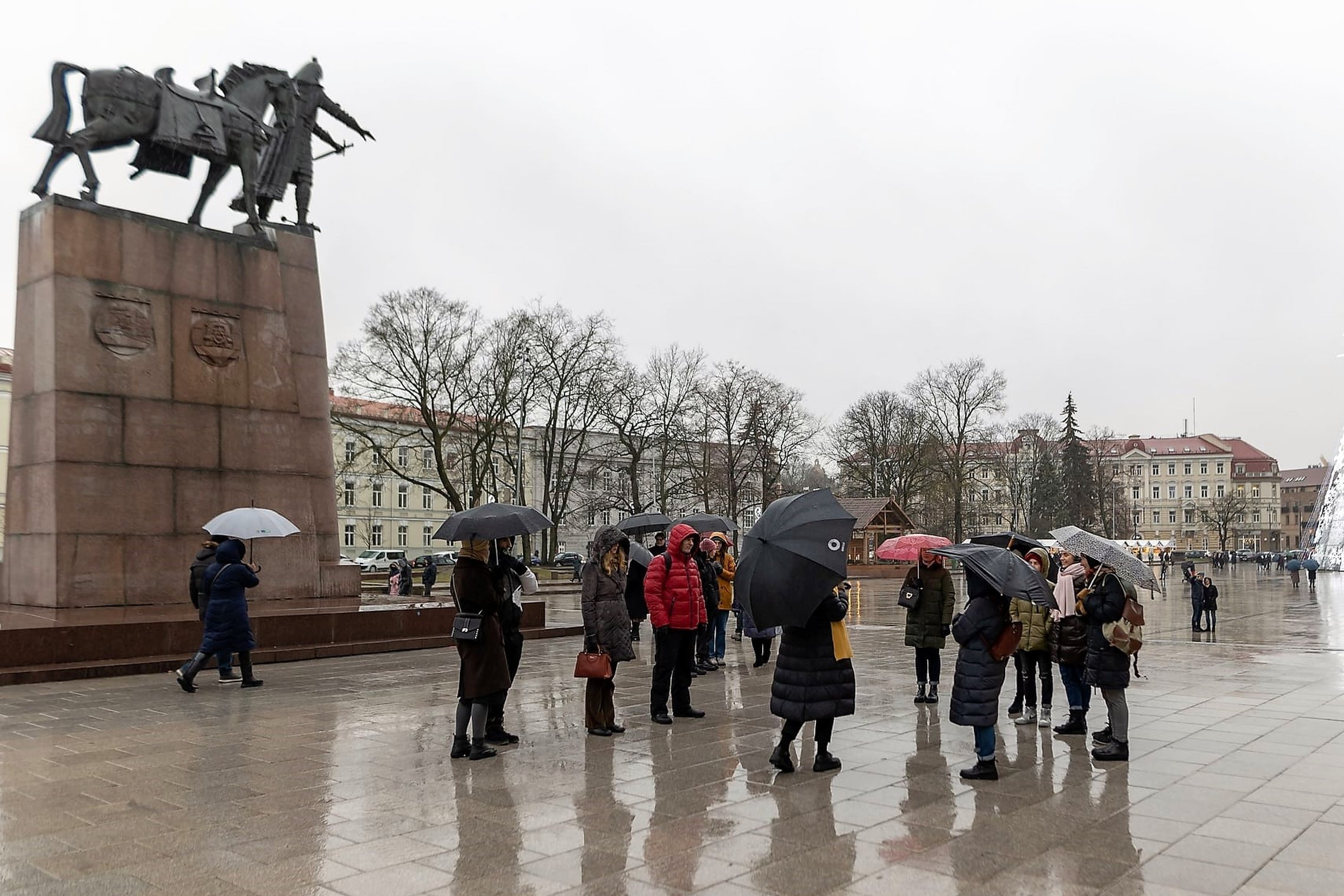 Būrelis žmonių renkasi ekskursijai Vilniaus katedros aikštėje prie Gedimino paminklo. Jie visi su skėčiais - pliaupia lietus.