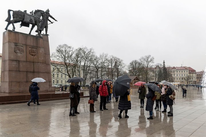 Būrelis žmonių renkasi ekskursijai Vilniaus katedros aikštėje prie Gedimino paminklo. Jie visi su skėčiais - pliaupia lietus. 
