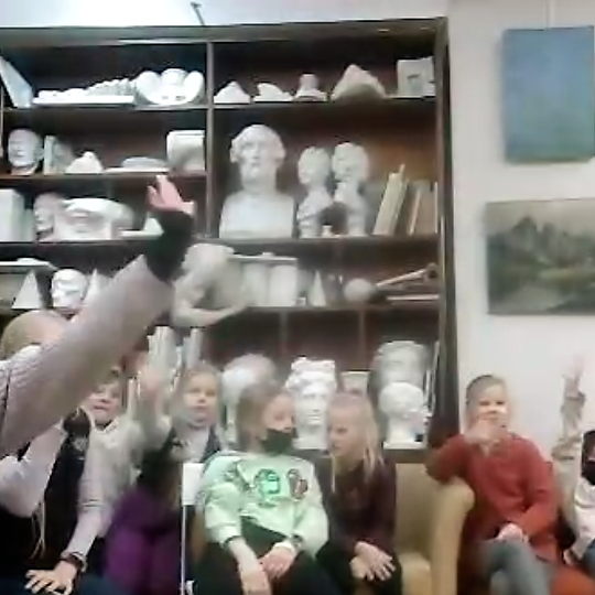 Dailės studija pilna gipsinių antikinių dievų galvų. Vaikų būrys sėdi ant sofos. Jie klausosi nuotolinės edukacijos, žiūrėdami į kompiuterio ekraną. Kai kuire vaikai yra su veido medicininėmis kaukėmis.