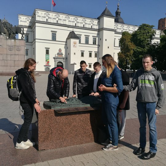 Kelių žmonių grupė stovi Vilniaus pilių rezervato teritorijoje. Jie apžiūrinėja čia esantį bronzinį Vilniaus pilių teritorijos maketą. Už jų nugarų yra balti valdovų rūmai ir senas Pilių parkas.