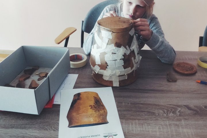 Nuotraukoje galima matyti 11 metų berniuką, sėdintį prie stalo. Ant stalo matome kartoninę dėžutę su keliomis likusiomis puodynės šukėmis. Šalia dėžės galima matyti lipnios juostos bei puodynės nuotrauka, kaip ji turėtų atrodyti. Ant lapo su puodynės pav
