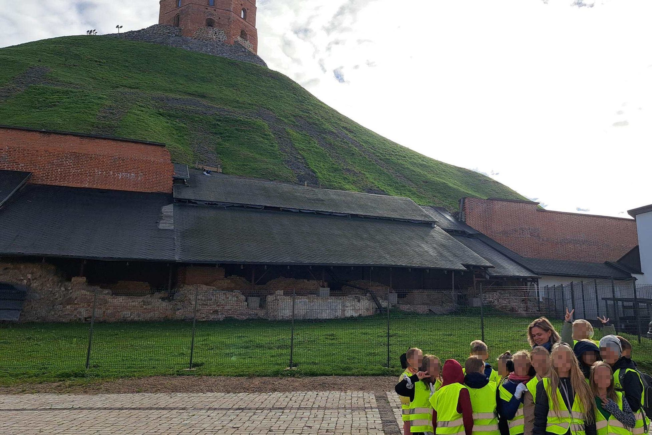 Mokiniai su ryškiaspalvėmis liemenėmis stovi Vilniaus pilių teritorijoje, už jų matyti Gedimino kalnas su Gedimino bokštu.
