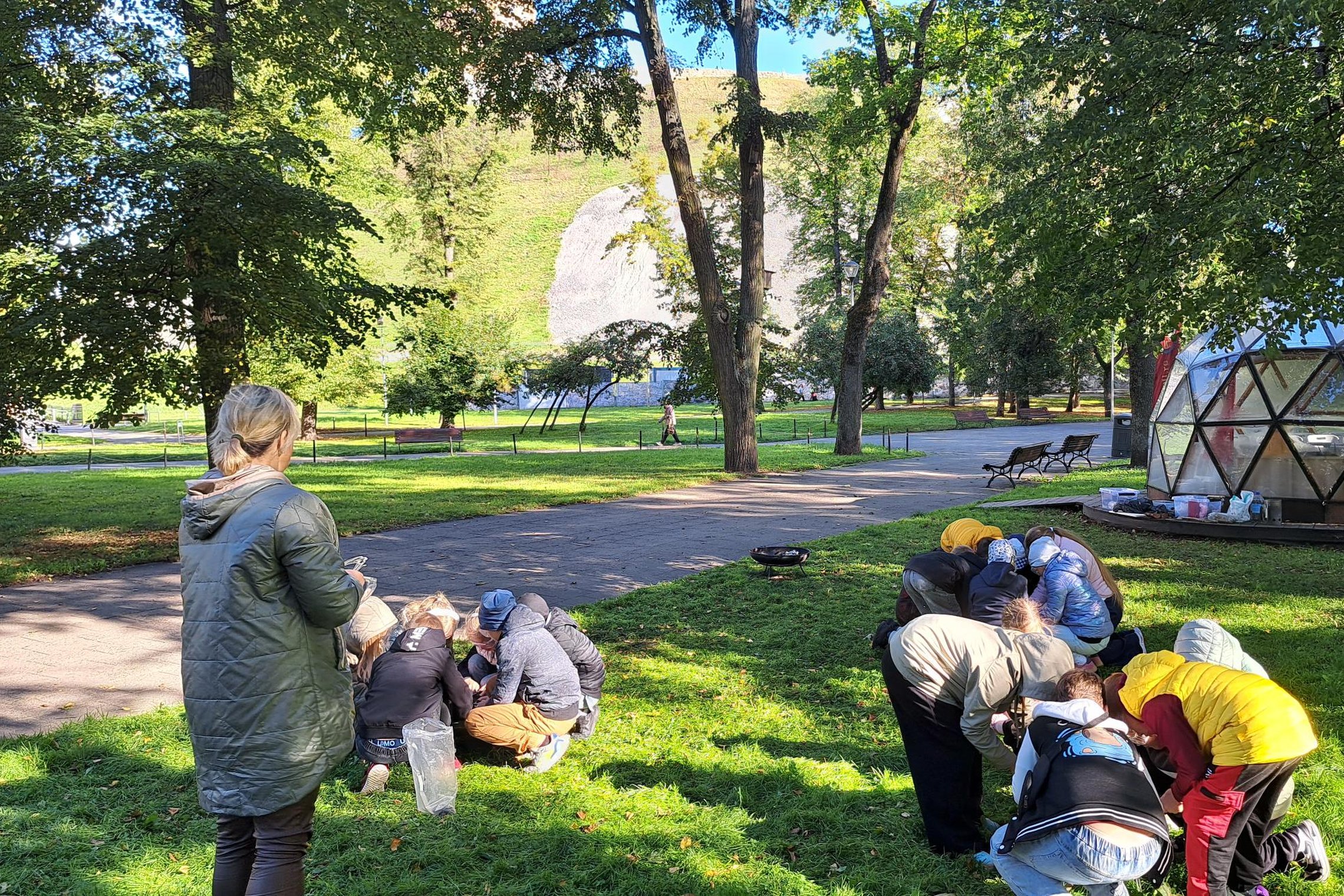 Vaikai, susibūrę į grupes, lauke prie Vilniaus pilių kupolo atlieka edukacinę užduotį apie ugnį, šalia stovi juos prižiūrinti mokytoja.