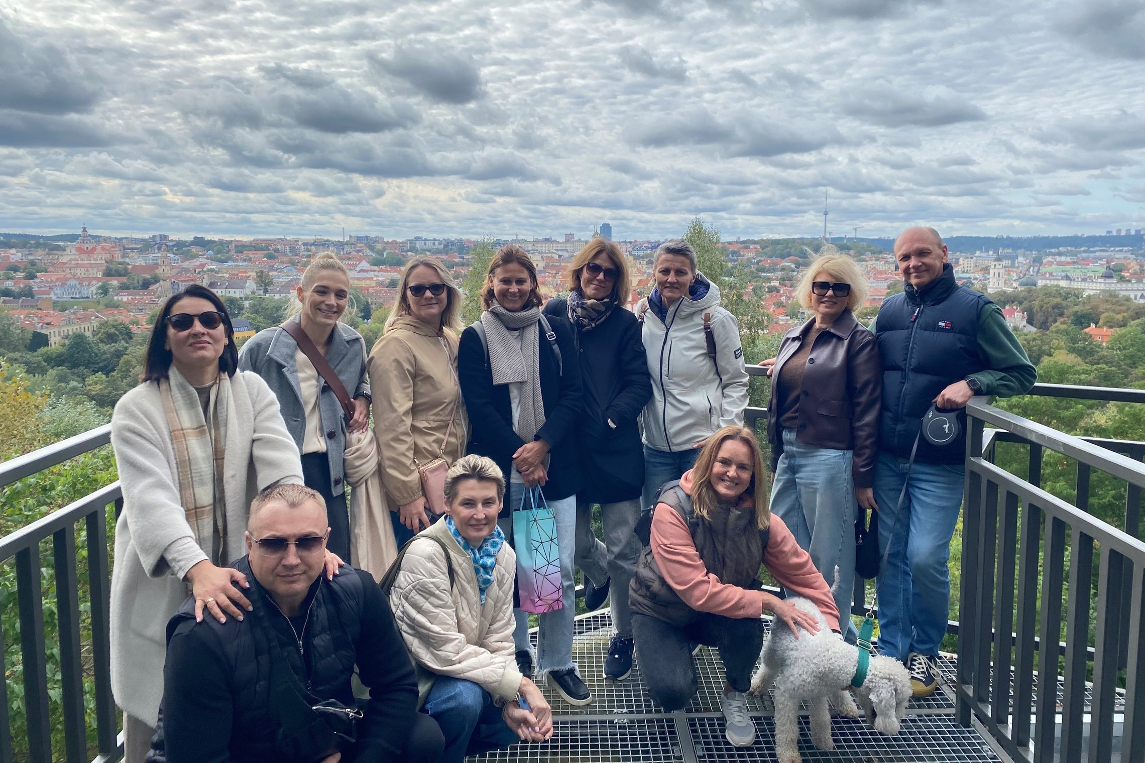 Grupė žmonių su šunimi pozuoja apžvalgos aikštelėje, fone – Vilniaus panorama debesuotu oru