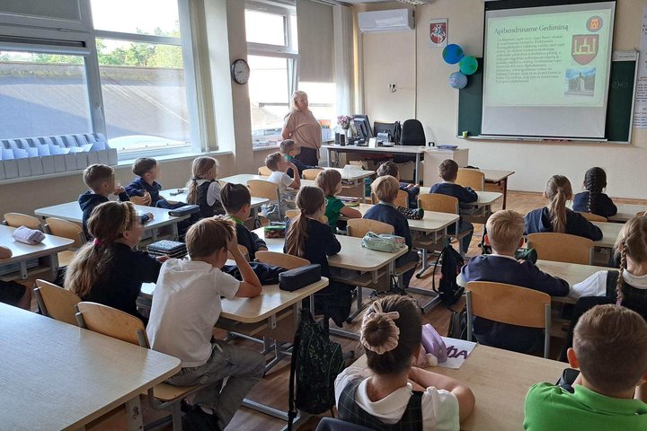Klasėje sėdintys pradinukai klausosi edukacijos apie Lietuvos didžiuosius kunigaikščius, priekyje stovi mokytoja ir rodomas skaidrių pristatymas apie Gediminą.
