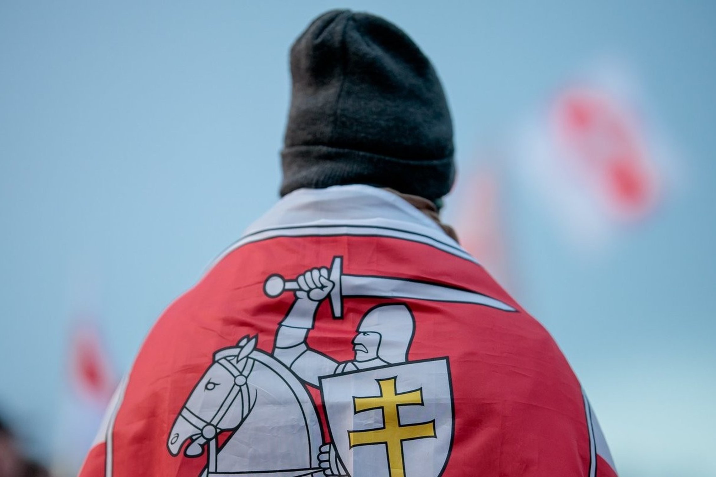 Asmuo su kepure apsigaubęs vėliava su raitelio ir kalaviją laikančio kario simboliu bei dvigubu kryžiumi.