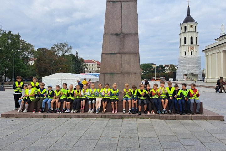Nuotraukoje didelė vaikų grupė ploja rankomis ir šypsosi, susėdę prie paminklo valdovui Gediminui.