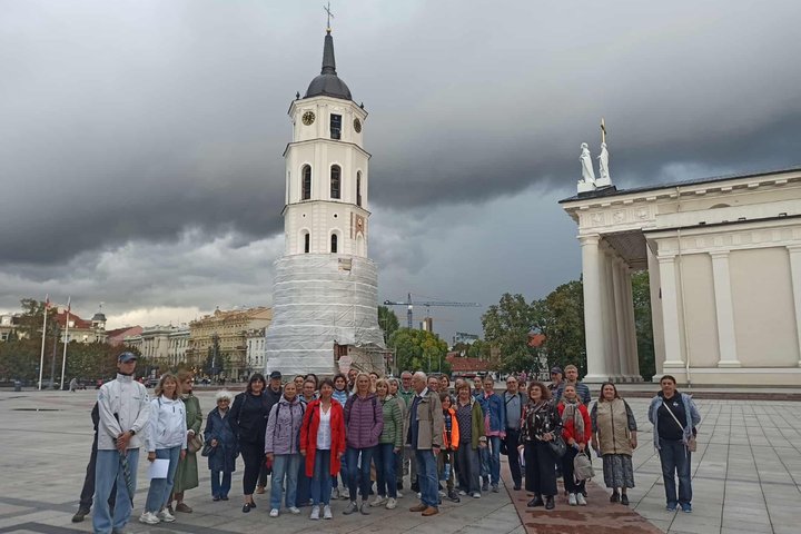 Nuotraukoje ekskursijos dalyviai pozuoja į kamerą. Už jų nugaros matosi varpinė.