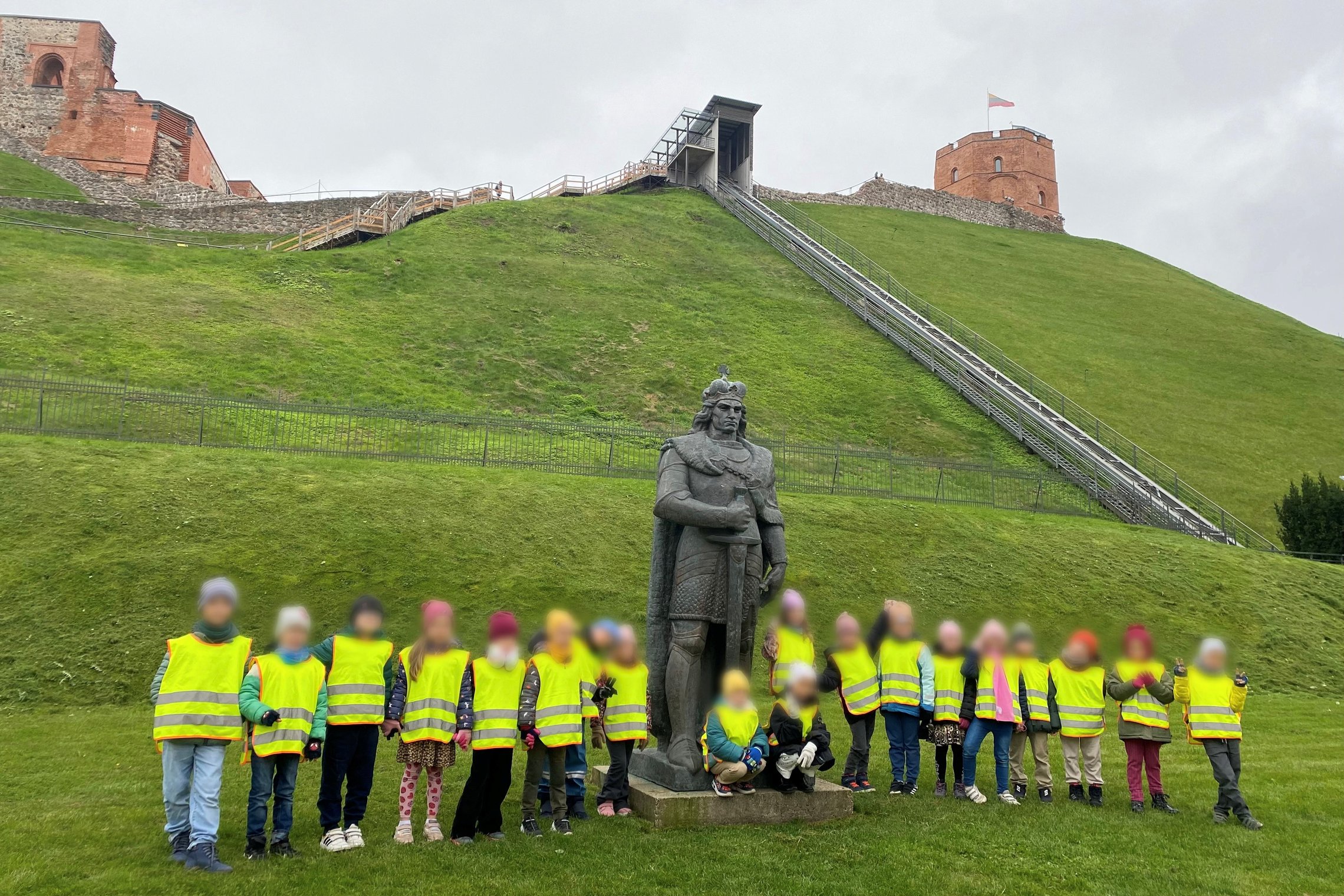 VDU licėjaus „Sokratus“ mokiniai su geltonomis saugos liemenėmis stovi prie Gedimino paminklo, už jų matosi Gedimino pilies bokštas ant žalio kalno su funikulieriumi