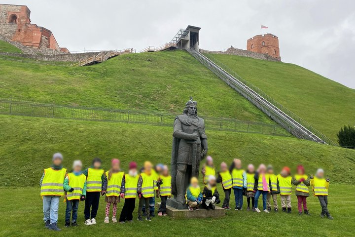 VDU licėjaus „Sokratus“ mokiniai su geltonomis saugos liemenėmis stovi prie Gedimino paminklo, už jų matosi Gedimino pilies bokštas ant žalio kalno su funikulieriumi