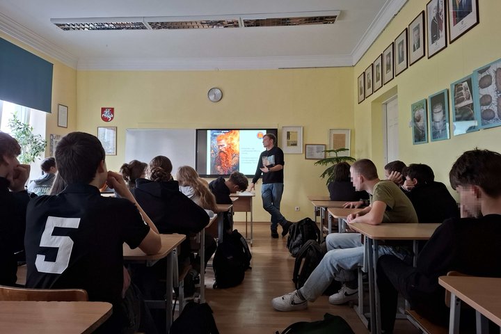 Klasėje sėdintys devintokai ir dešimtokai klausosi edukatoriaus pasakojimo apie baltų tikėjimą; ekrane rodoma Perkūno iliustracija.