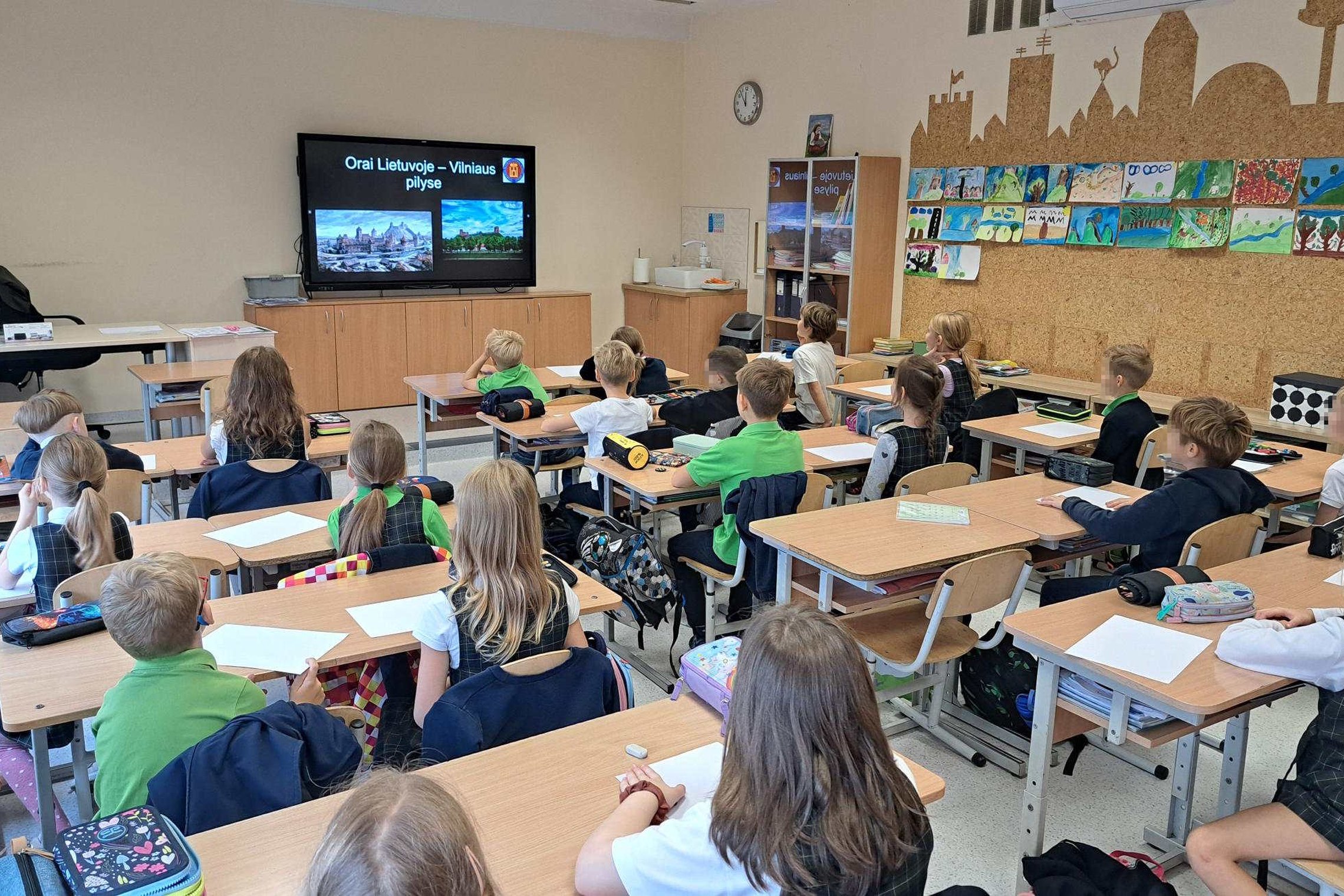 Antrokai klasėje sėdi prie savo suolų ir žiūri į ekraną, kuriame rodoma skaidrė „Orai Lietuvoje – Vilniaus pilyse