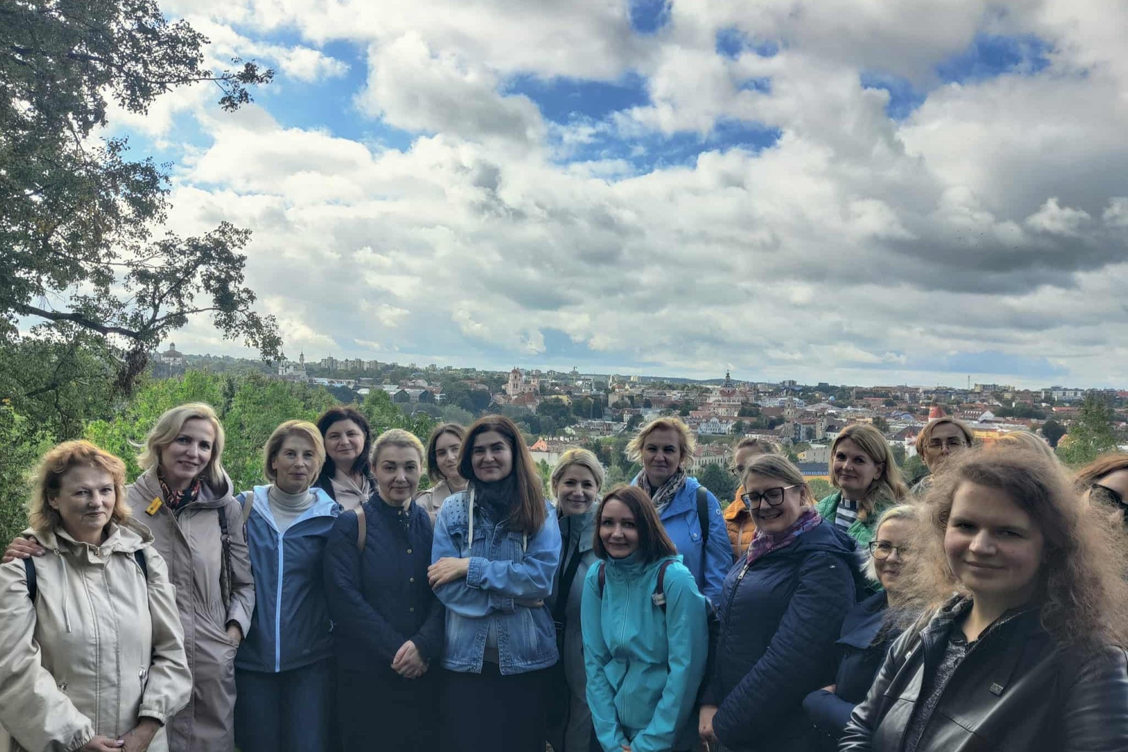 Vilniaus pilių direkcijos kolektyvas stovi ant Altanos kalno su Vilniaus senamiesčio panorama fone, matosi bažnyčių bokštai ir raudoni stogai po debesuotu dangumi.