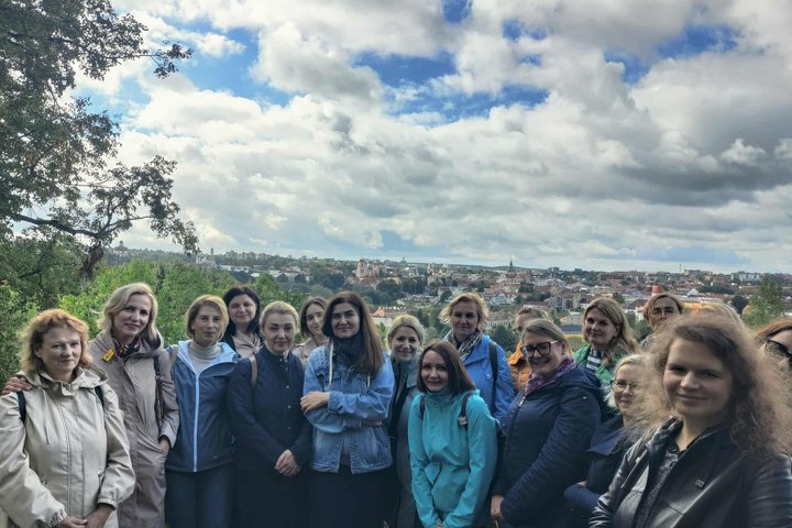 Vilniaus pilių direkcijos kolektyvas stovi ant Altanos kalno su Vilniaus senamiesčio panorama fone, matosi bažnyčių bokštai ir raudoni stogai po debesuotu dangumi.