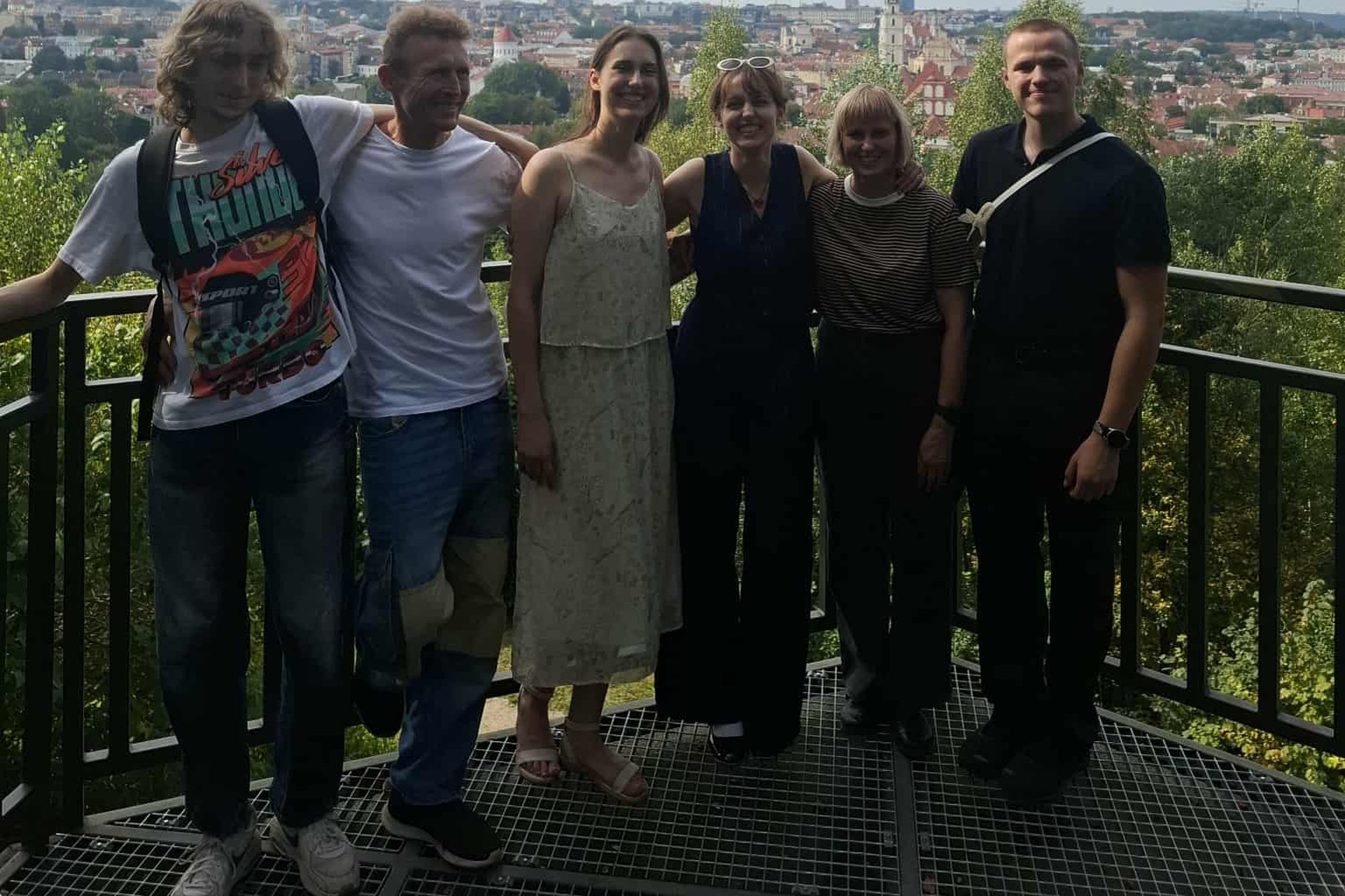 Šeši žmonės stovi ant altanos bokšto aikštelės Vilniaus pilių teritorijoje, už jų atsiveria miesto panorama su bažnyčių bokštais ir televizijos bokštu tolumoje.