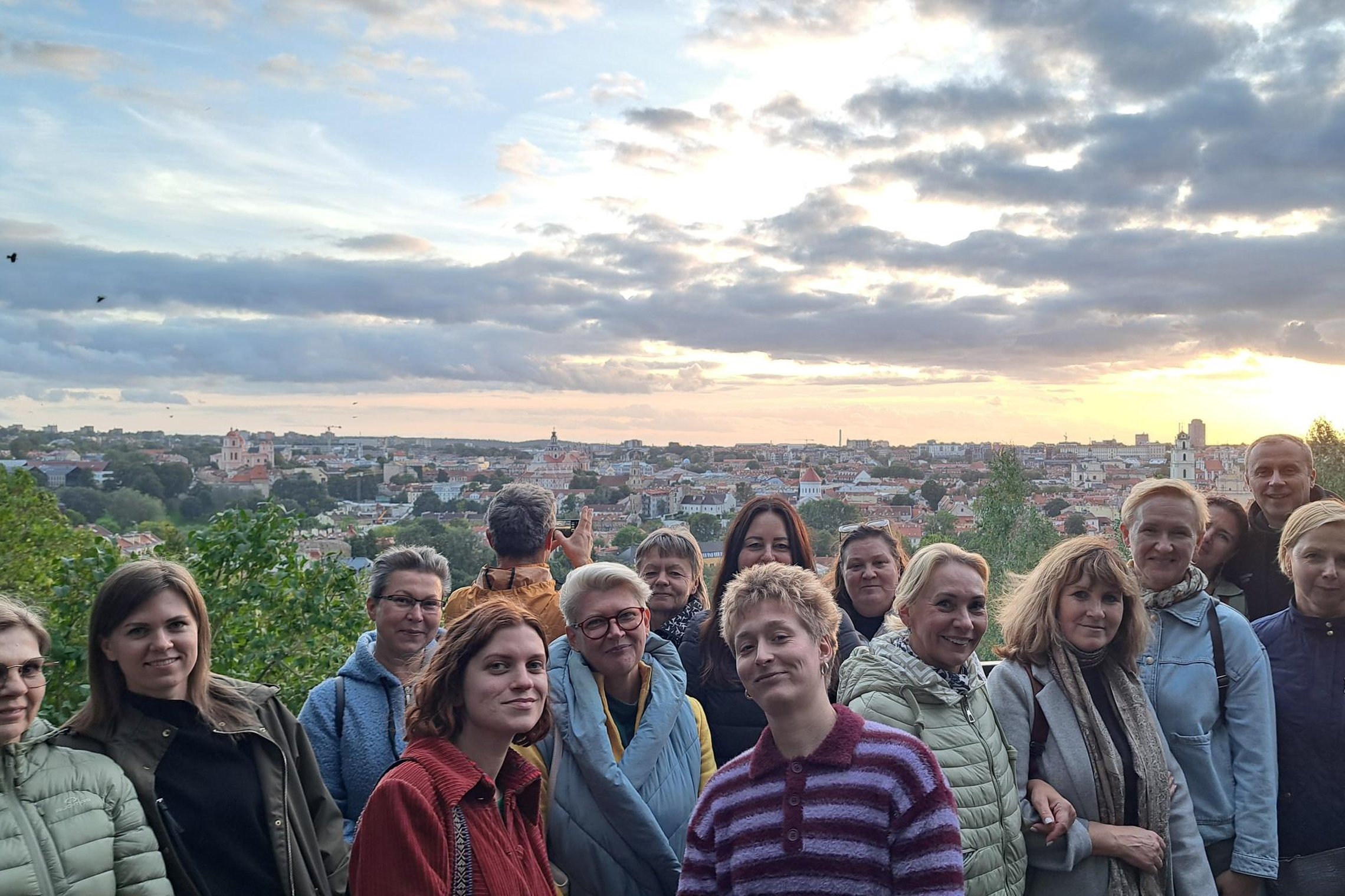 Grupė žmonių stovi Vilniaus pilių teritorijoje ant altanos aikštelės, už jų matyti Vilniaus senamiesčio panorama ir saulėlydis.