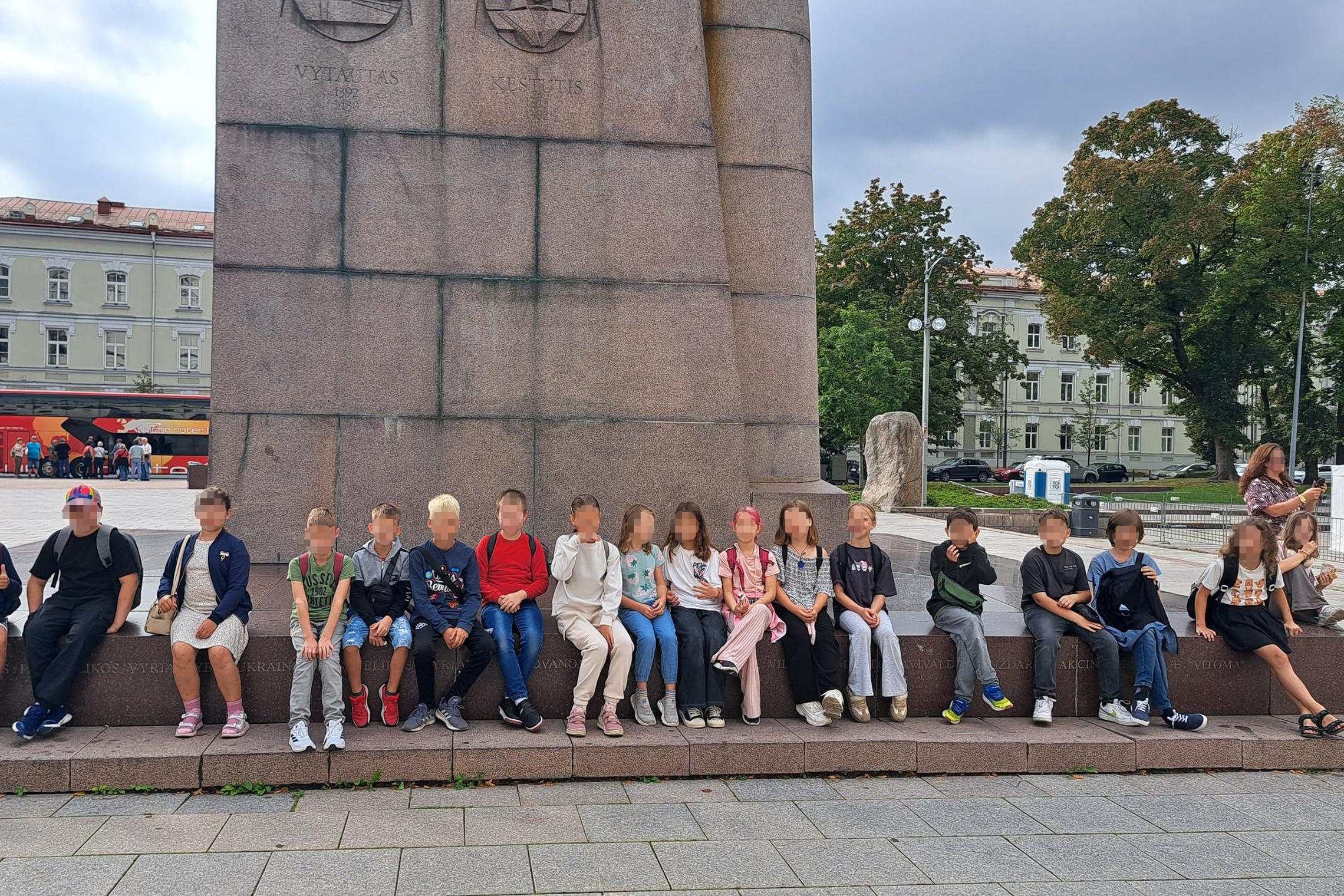 Vaikų grupė sėdi prie paminklo Vilniaus Katedros aikštėje, už jų matosi senamiesčio pastatai ir raudonas turistinis autobusas.