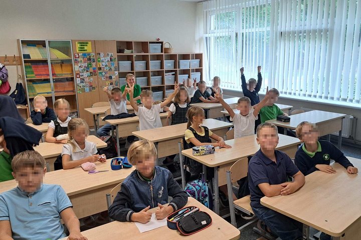 Trečiokų klasė per edukaciją apie Mindaugą ir Gediminą – mokiniai sėdi už suolų, kai kurie kelia rankas ar rodo pergalės ženklus, klasėje šviesu ir jauku.