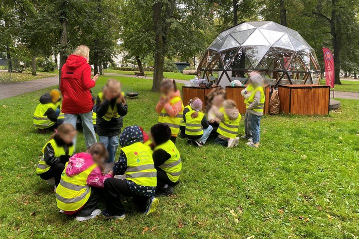 Vaikai su geltonomis liemenėmis, susibūrę grupelėmis ant žolės prie kupolo formos pastato, mokosi senovinių ugnies kūrimo būdų edukacijos metu