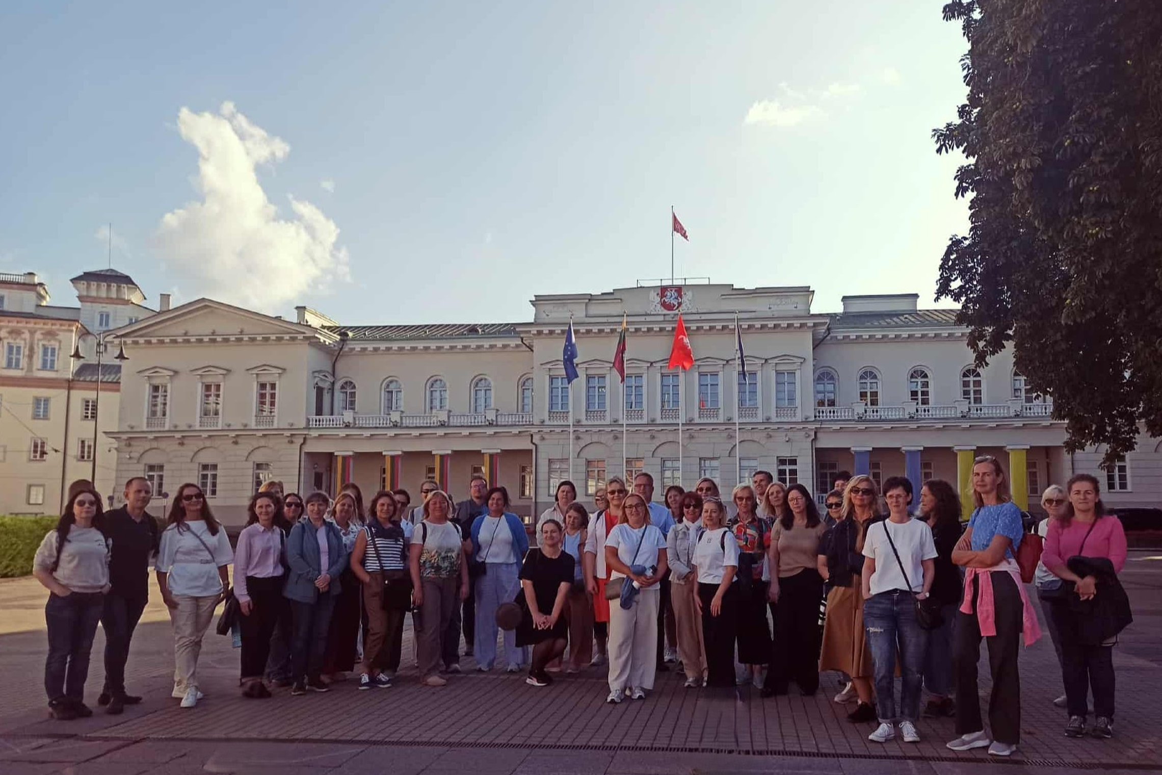 Nuotraukoje didelė žmonių grupė pozuoja ir šypsosi. Jie dalyvauja ekskursijoje.