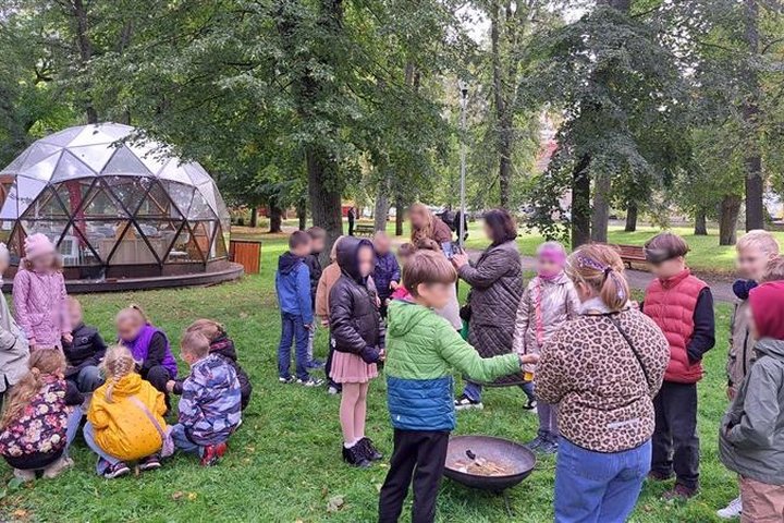 Nuotraukoje vaikai, pasiskirstę į tris grupeles, bando įžiebti kibirkštį su skeltuvais. Vieni dirba pasilenkę, kiti dairosi ir mokosi iš draugų.