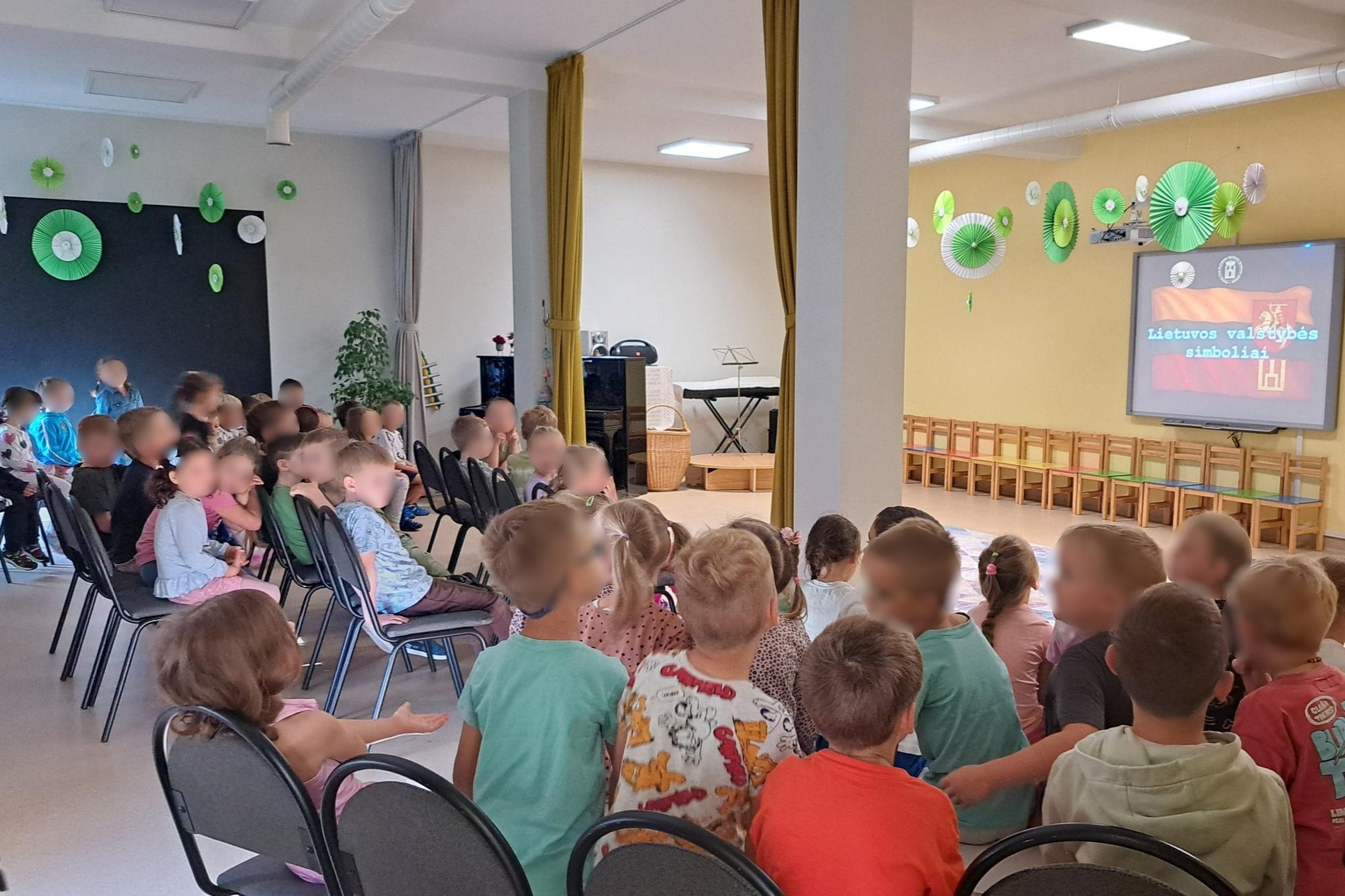 Nuotraukoje salė su ekranu ir trys grupės vaikų dalyvauja edukacijoje apie valstybės simbolius.