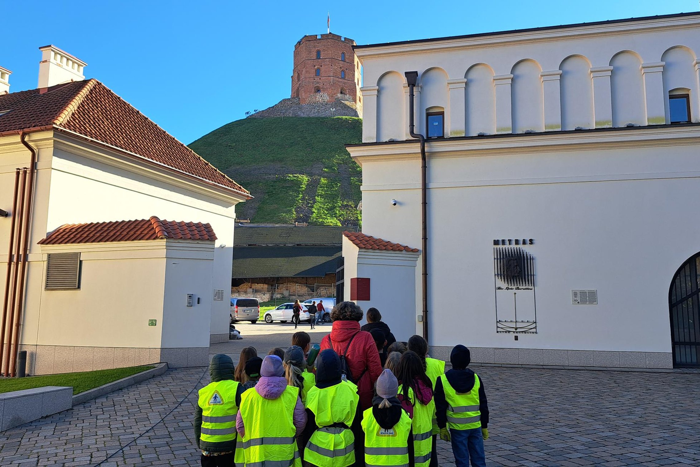 Vaikų grupė su ryškiai geltonomis liemenėmis ir lydinčiu mokytoju stovi Vilniaus pilių teritorijoje, žvelgdami į Gedimino pilies bokštą ant kalno.