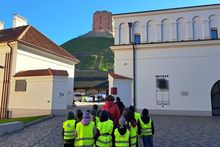 Vaikų grupė su ryškiai geltonomis liemenėmis ir lydinčiu mokytoju stovi Vilniaus pilių teritorijoje, žvelgdami į Gedimino pilies bokštą ant kalno.