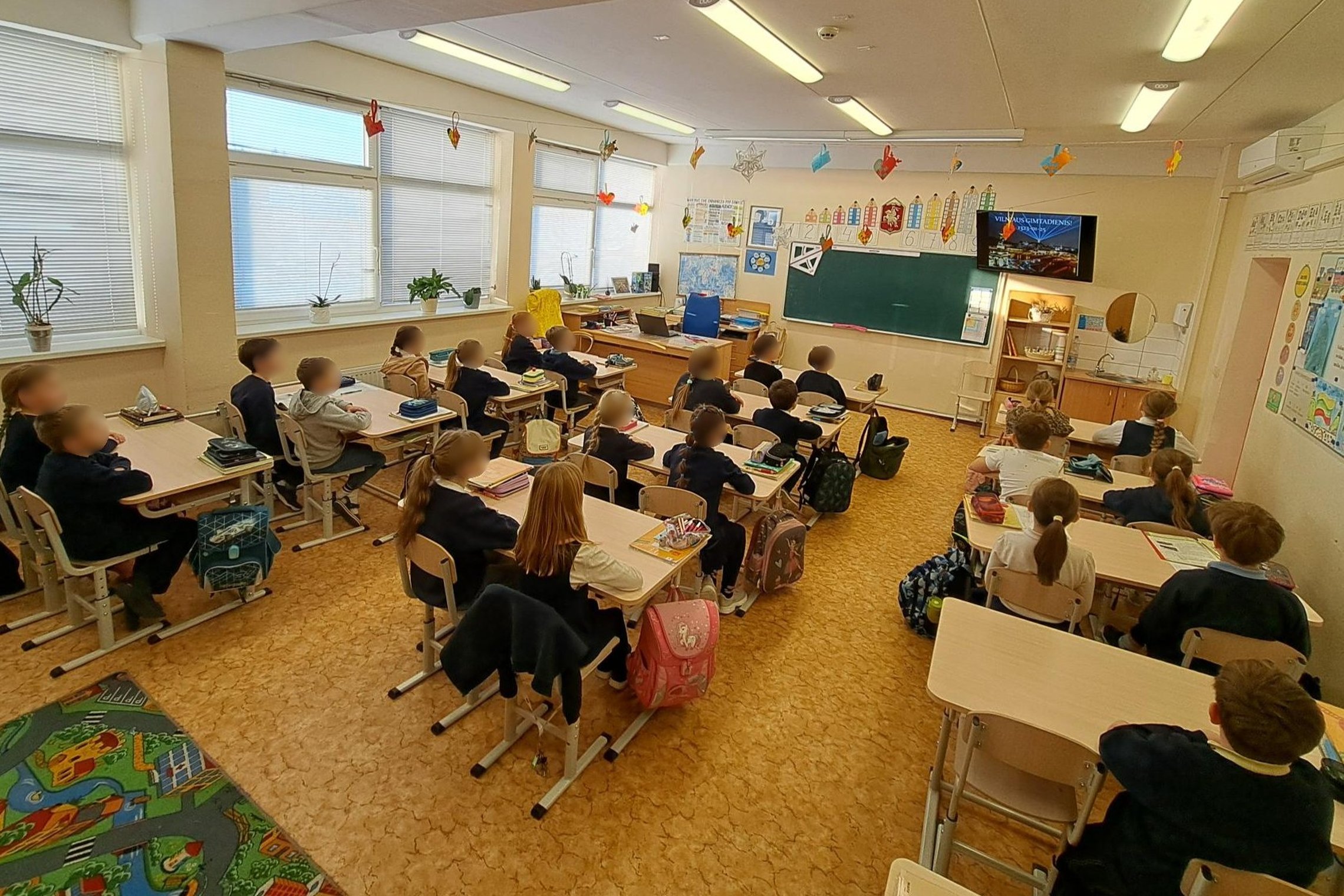 Nuotraukoje vaikai sėdi suoluose. Žiūri į ekraną klasės priekyje. Ekrane edukacijos pavadinimas „Vilniaus gimtadienis ir kunigaikštis Gediminas“