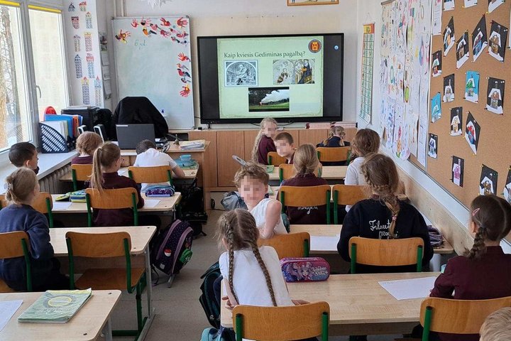 Klasėje prie suolų sėdintys pradinės mokyklos pirmokai dalyvauja edukacijoje – vaikai klausosi pasakojimo ir ekrane rodomos medžiagos apie gyvenimą praeityje.