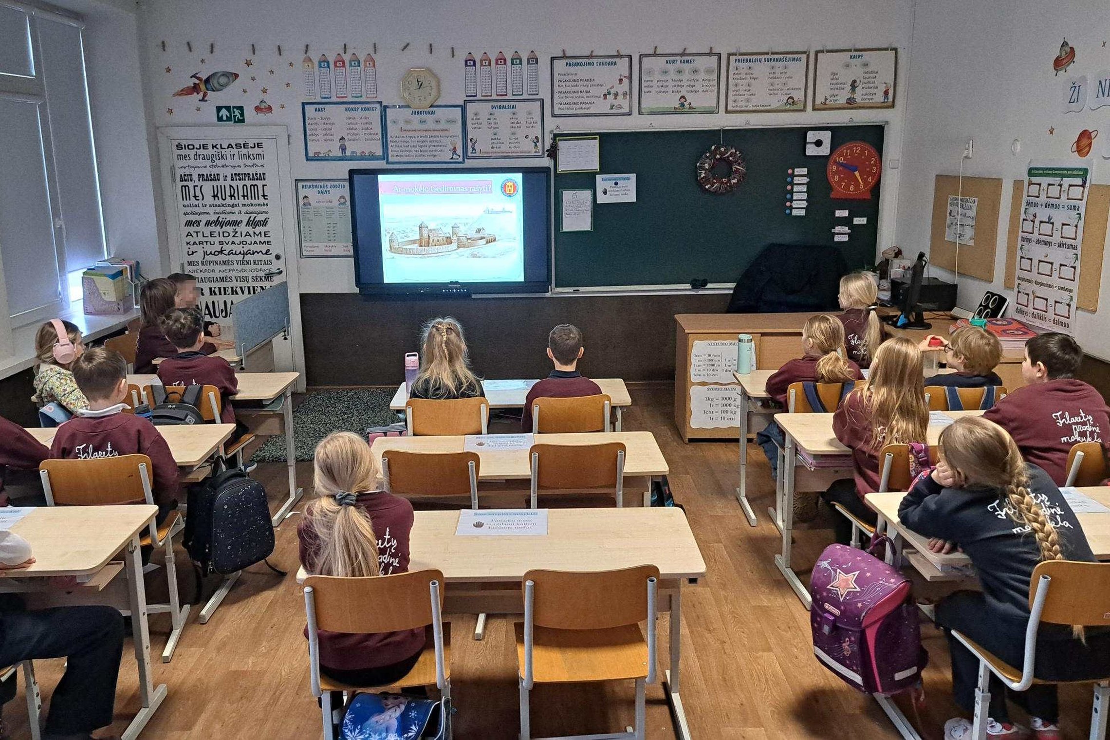 Klasėje prie suolų sėdi Vilniaus Filaretų pradinės mokyklos mokiniai, stebintys ekrane rodomą pristatymą apie Lietuvos didžiuosius kunigaikščius ir Vilniaus pilis.