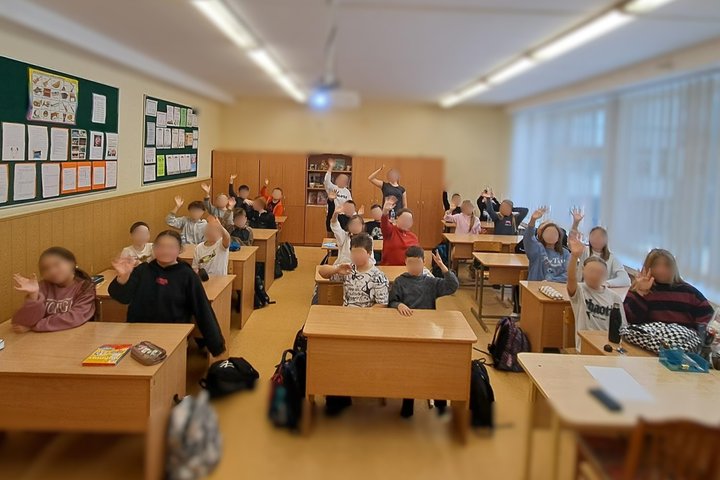Nuotraukoje klasė pilna vaikų. Vaikai sėdi suoluose ir mojuoja. Atrodo patenkinti ir linksmi, galimai jiems patiko edukacija.