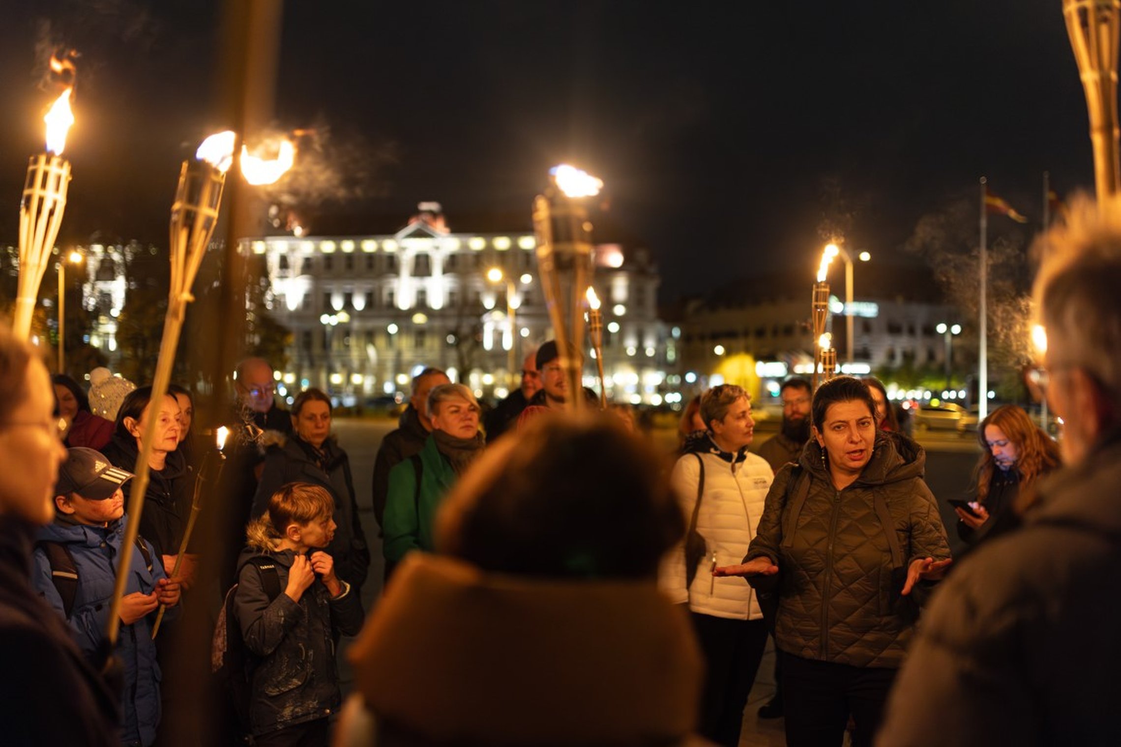 Vakarinės ekskursijos dalyviai miesto aikštėje klausosi gidės, laikydami degančius fakelus. Fone matyti apšviesti miesto pastatai ir naktinė miesto atmosfera.