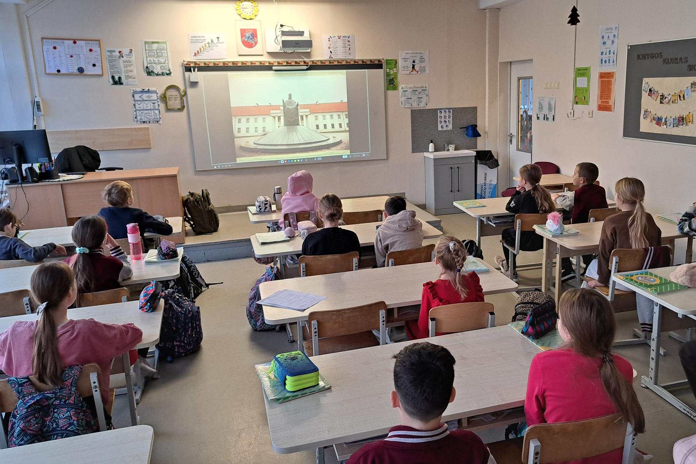 klasėje sėdintys trečios klasės mokiniai klausosi edukacijos apie Lietuvos valstybės sukūrimą; priekyje matomas projektorius su istoriniu vaizdu, vaikai sėdi suoluose ir žiūri į ekraną.